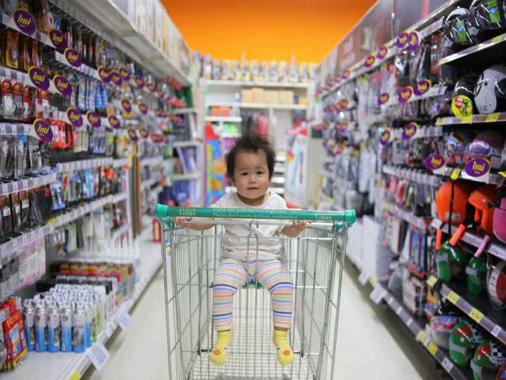 Baby sitting on a shopping cart in the middle of a supermarket aisle.