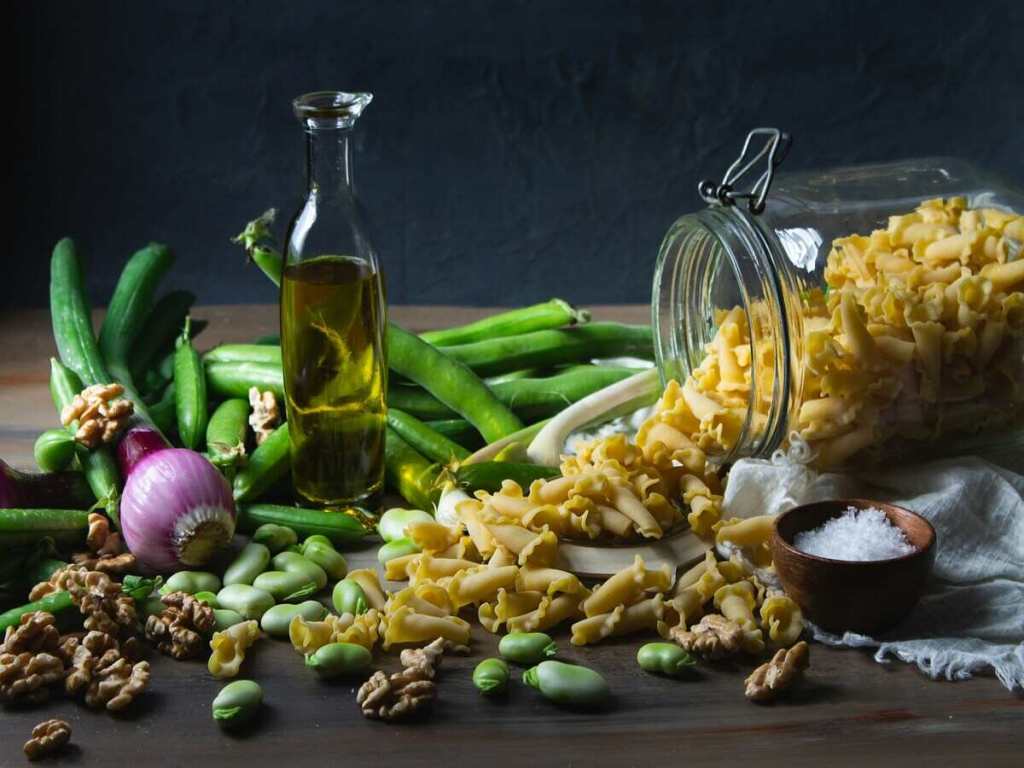 A glass bottle of olive oil surrounded by pasta, nuts and vegetables.