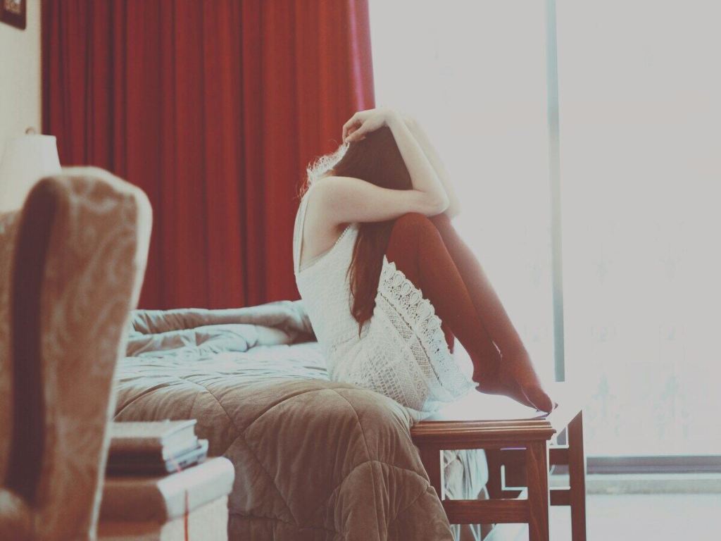A woman sitting on the bed and hiding her face with her hands.