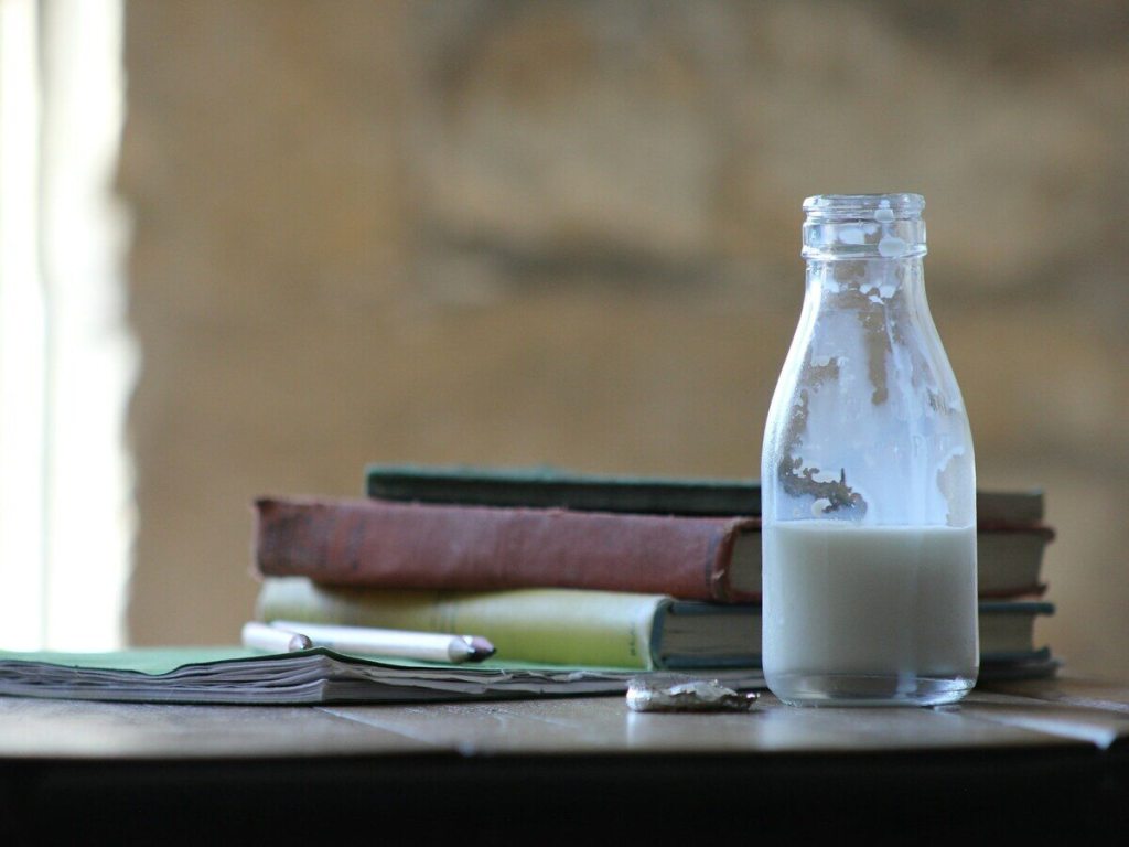 A glass bottle half filled with milk on a desk with notebooks.