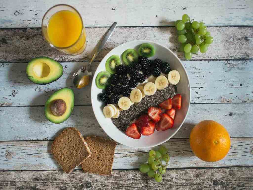 A healthy bowl of kiwi, banana, strawberries, blackberries with avocadoes, brown bread, grapes and oranges on the side.
