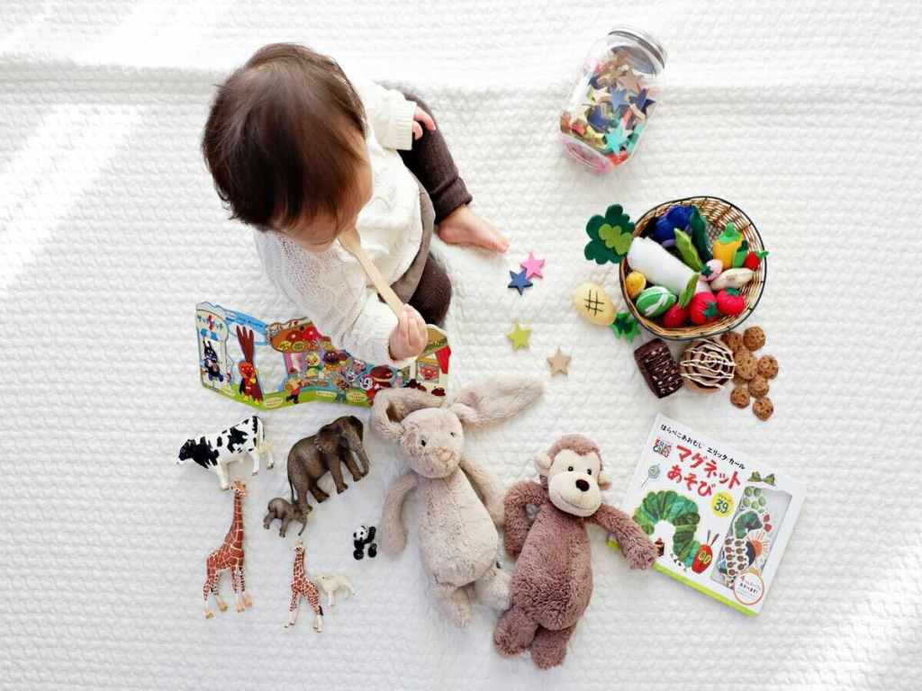 A baby is sitting on the floor and surrounded by various children's toys, including stuffed and plastic animals.